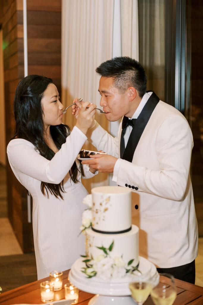 A couple in formal attire shares a piece of cake. A two-tier cake and lit candles are in the foreground. They are indoors.
