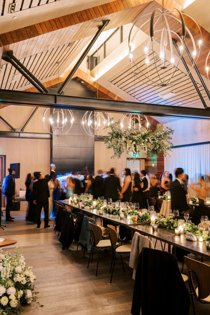 A large dining room with long tables set for a formal event. People are mingling under modern chandeliers and a high wooden ceiling.