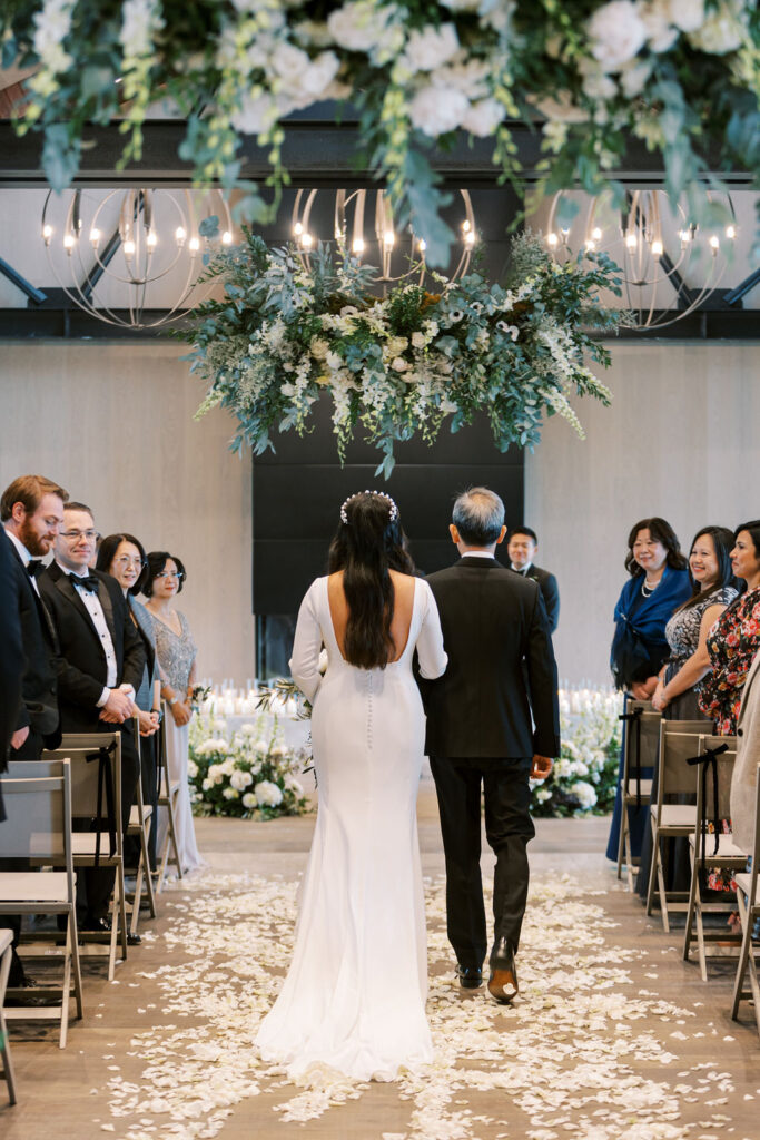 A bride and an older man walk down an aisle covered with white petals, surrounded by seated guests, under a large floral chandelier.