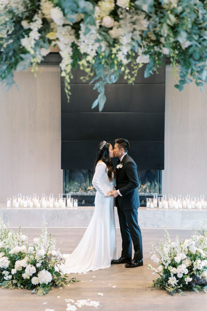 A bride and groom kiss at their wedding ceremony, framed by floral arrangements and white candles.