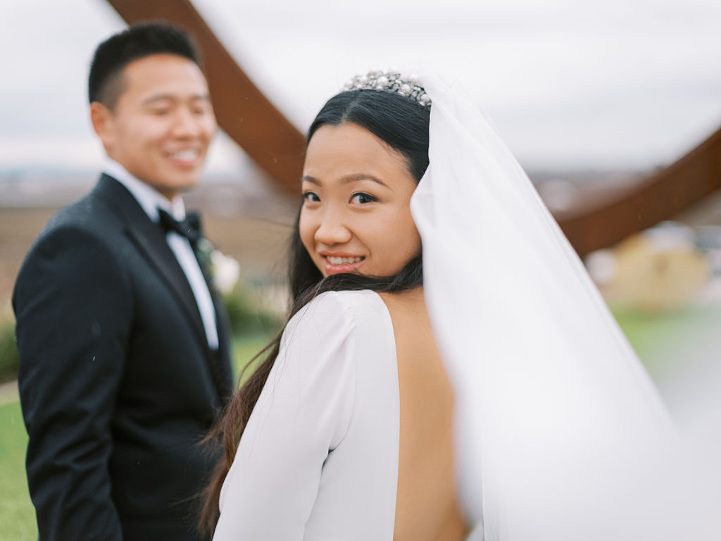 Bride in a white dress smiles at the camera with a groom in a black suit in the background. They are outdoors with a blurred, natural setting.