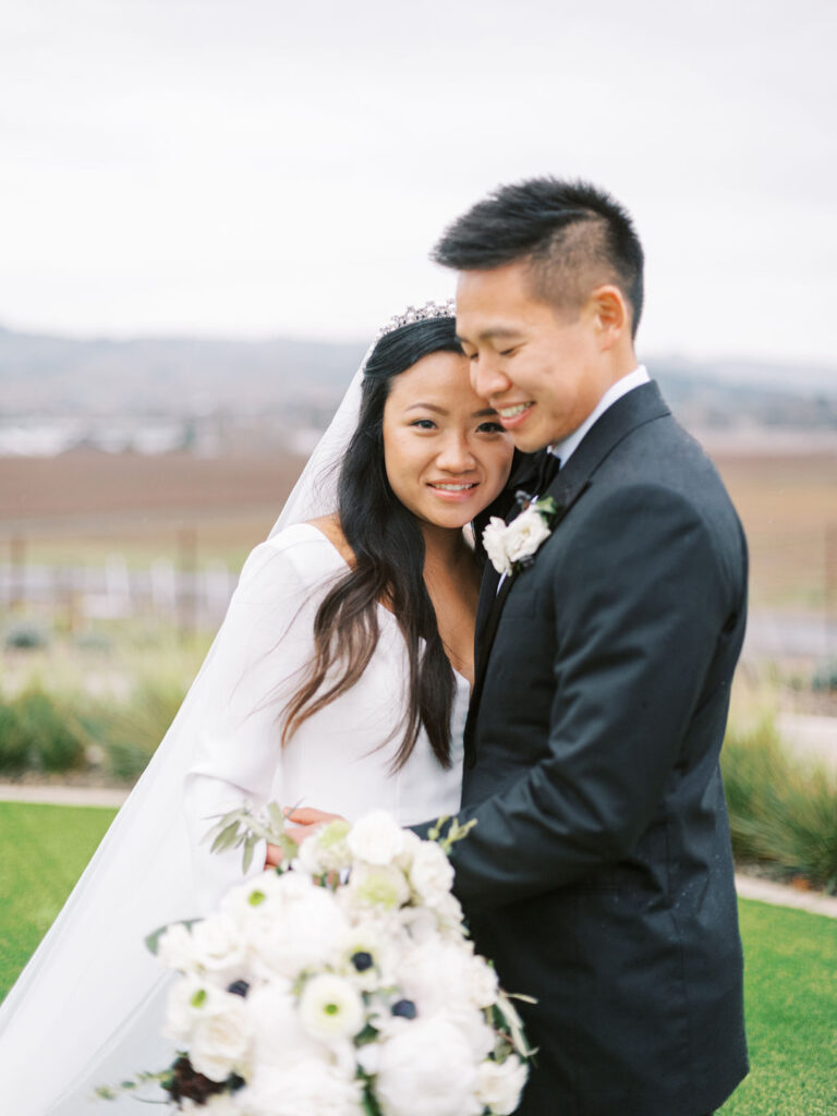 A couple in wedding attire stands outdoors. The bride holds a bouquet of white flowers. The background features grass and distant hills under a cloudy sky.