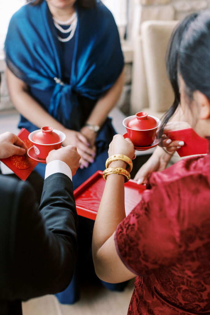 A woman and man hold red tea cups and red envelopes, participating in a traditional tea ceremony. A seated woman in a blue shawl watches.