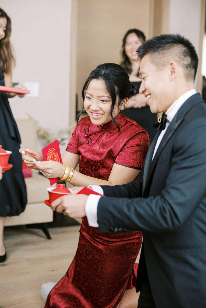 A couple in traditional attire is kneeling, holding red envelopes and teacups, surrounded by others during a tea ceremony.