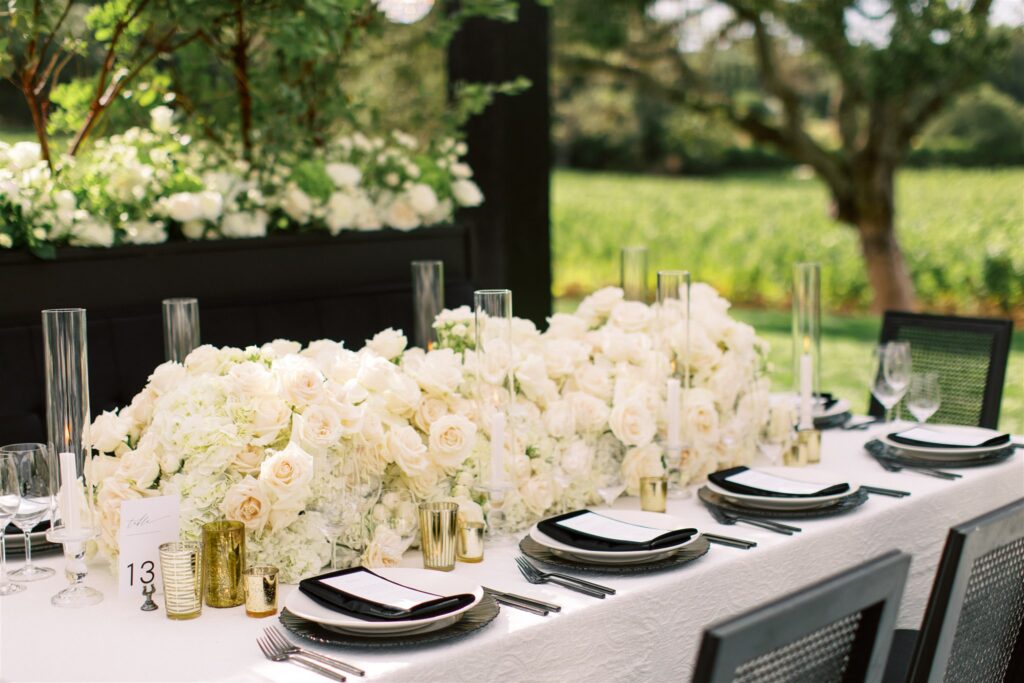 Elegant outdoor dining table with a white tablecloth, black plates, gold accents, and a floral centerpiece of white roses and hydrangeas, set beside a vineyard.