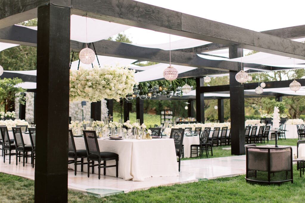 Elegant outdoor dining setup with black chairs, white tablecloths, and hanging crystal decorations under a black pergola. White floral centerpieces adorn the tables.