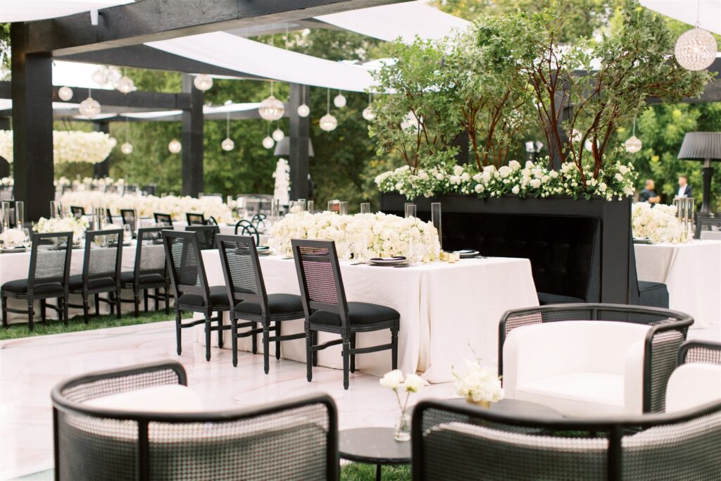 Elegant outdoor wedding reception setup with white tablecloths, black chairs, floral centerpieces, and overhead string lights.