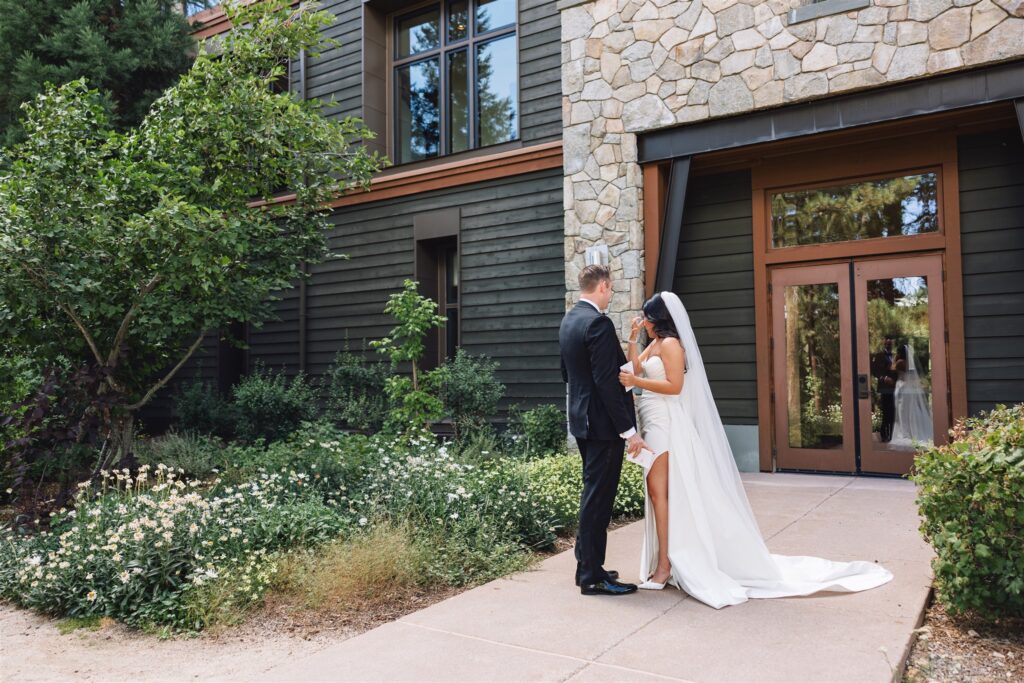 A couple in wedding attire stands holding hands in front of a building with stone and wood siding, surrounded by greenery and flowers.