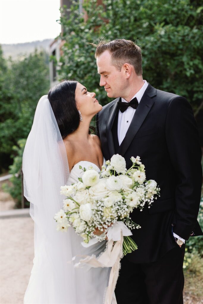 A bride and groom share a loving gaze outdoors. The bride holds a bouquet of white flowers and wears a veil. The groom is in a black suit and bowtie. Greenery and buildings are in the background.