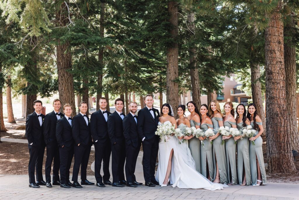 A wedding party poses in a wooded outdoor setting. The group includes bridesmaids in matching light green dresses and groomsmen in black suits.