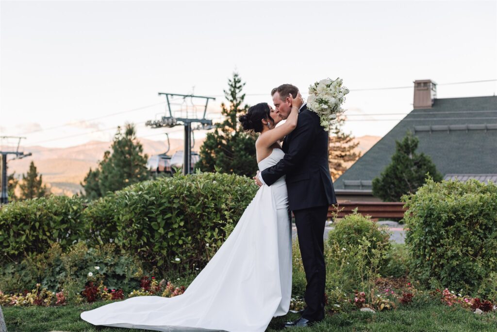 Bride and groom kissing in a garden setting with mountains and a lodge in the background.