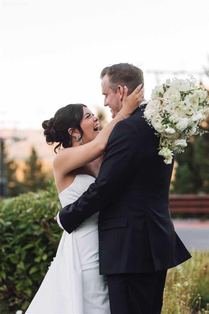 A bride and groom embrace outdoors, with the bride holding a bouquet of white flowers.