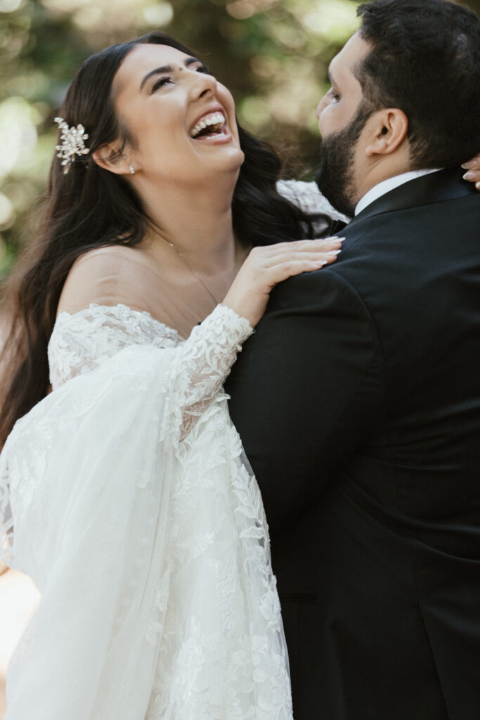 Bride and groom laughing together, outdoors. Bride in lace gown with off-shoulder design, groom in black suit. Lush greenery in the background.