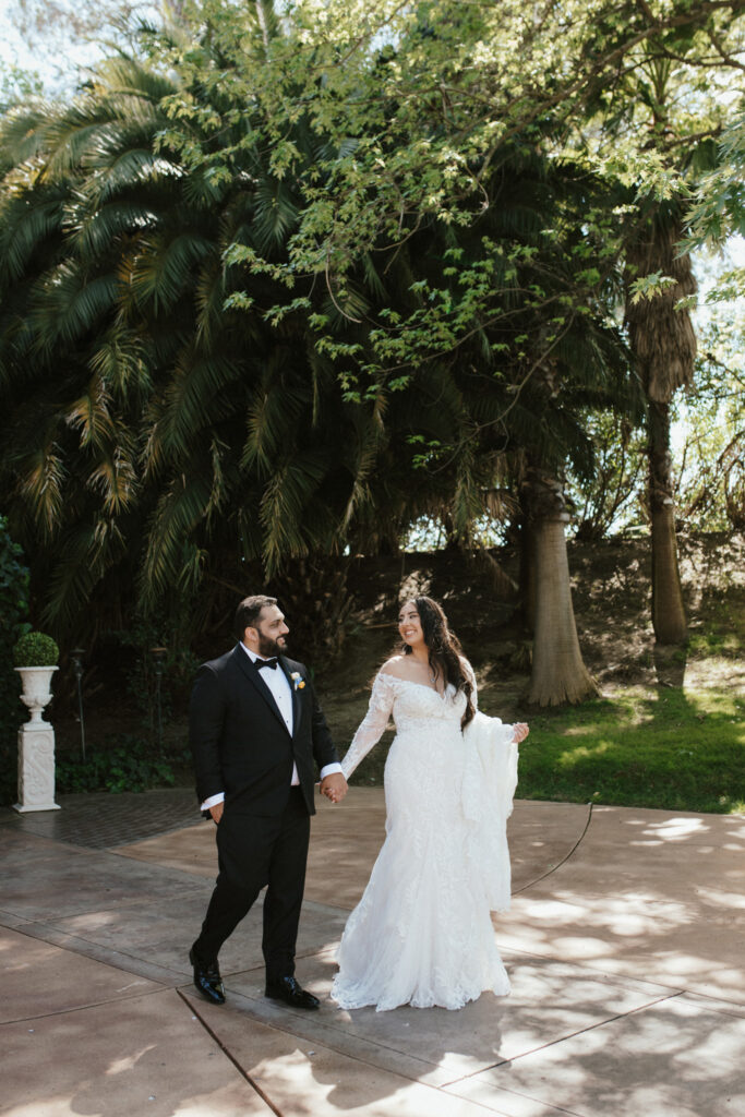 A bride and groom walk hand in hand outdoors, surrounded by lush greenery. The bride wears a white lace gown, and the groom is in a black tuxedo.