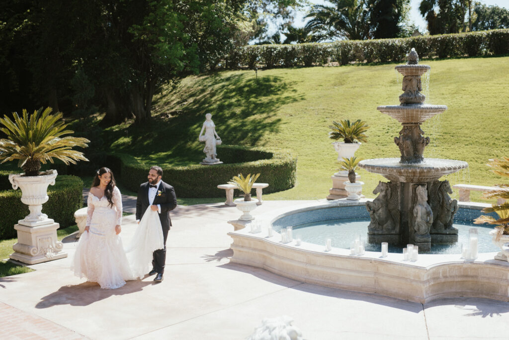 Bride and groom walk by a decorative fountain in a garden setting, surrounded by greenery and sculptures.