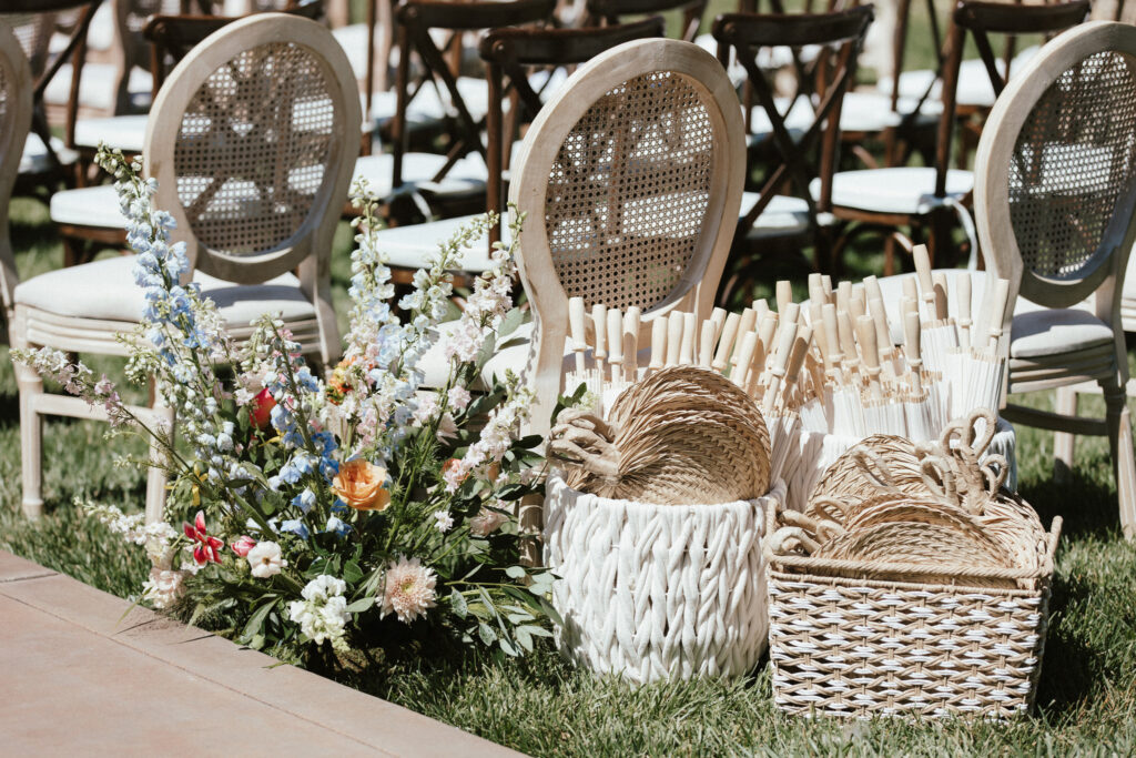 Wedding ceremony setup with wooden chairs on grass, floral arrangements, and baskets filled with woven fans and paper umbrellas.