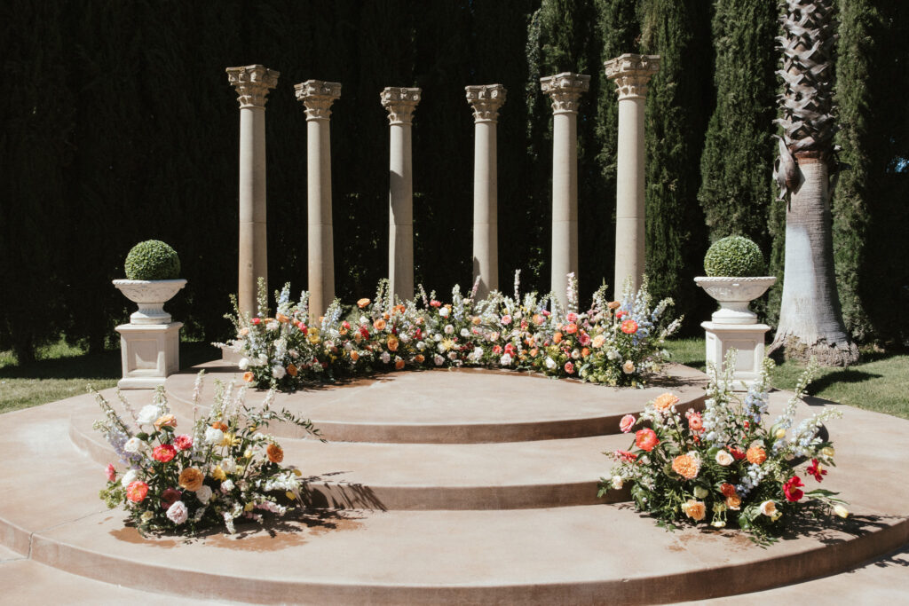 Outdoor wedding altar with a circular stone platform, decorated with colorful flower arrangements and flanked by tall columns.