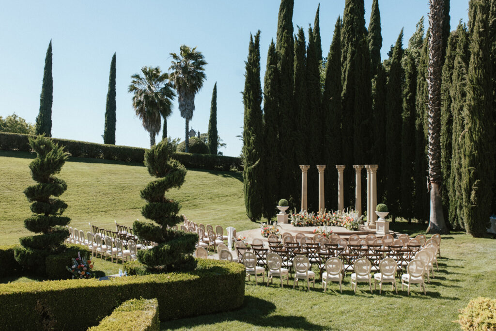 Outdoor wedding ceremony setup with chairs arranged in a semi-circle around a decorative altar, surrounded by tall trees and manicured hedges.