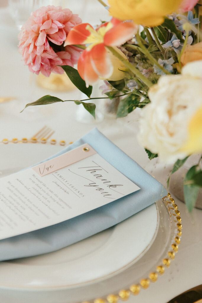 A floral centerpiece with pink and yellow flowers is next to a place setting, featuring a thank you card on a blue napkin atop a white plate with a decorative gold-rimmed edge.