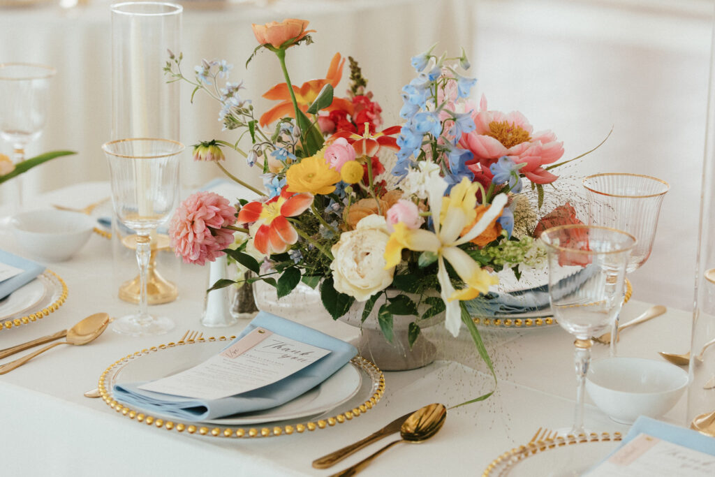 Elegant table setting with a colorful floral centerpiece, gold-rimmed glassware, and light blue napkins on white plates.