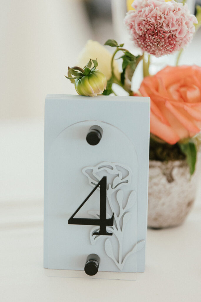 Table number four sign with floral cutout, set beside a small flower arrangement in a pot, including roses and other blooms.