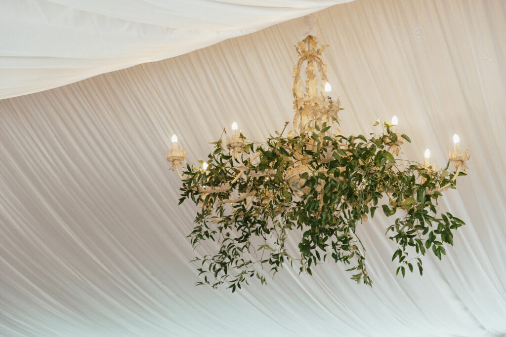 An ornate chandelier decorated with greenery hangs from a white draped ceiling.
