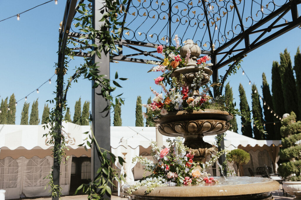 A stone fountain adorned with flowers under a metal pergola, surrounded by tents and trees, with string lights overhead.