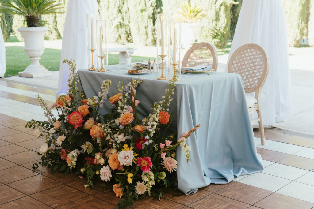A table with a light blue tablecloth is set with candles and plates, surrounded by an arrangement of colorful flowers.