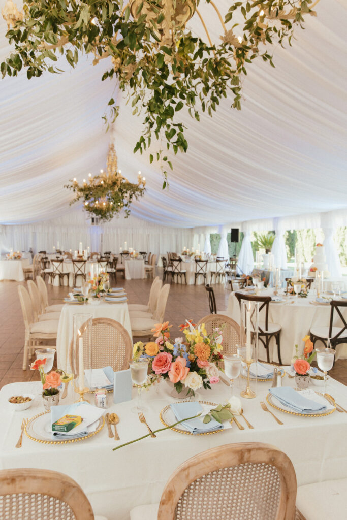 Elegant wedding reception setup under a white marquee with chandeliers, decorated tables with floral centerpieces, candles, and polished tableware.
