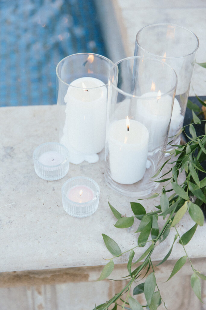Lit white candles in glass holders on a stone ledge, with green foliage, beside a blue-tiled pool.