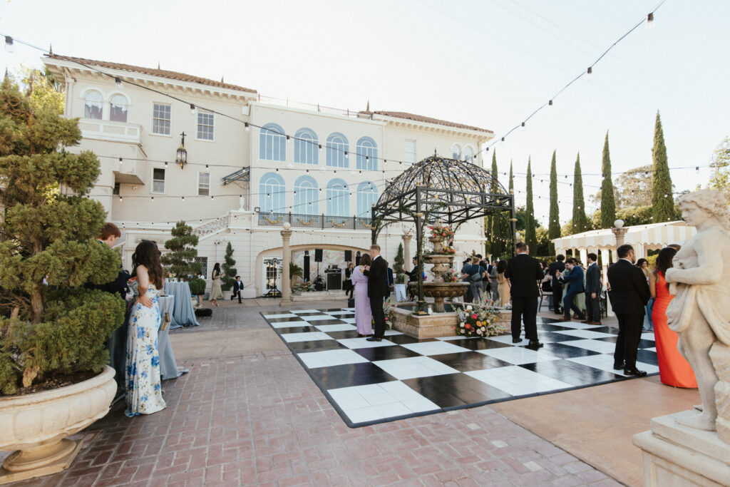 Outdoor gathering with people in formal attire around a black-and-white checkered dance floor, near a fountain, in front of a large building with arches and columns.