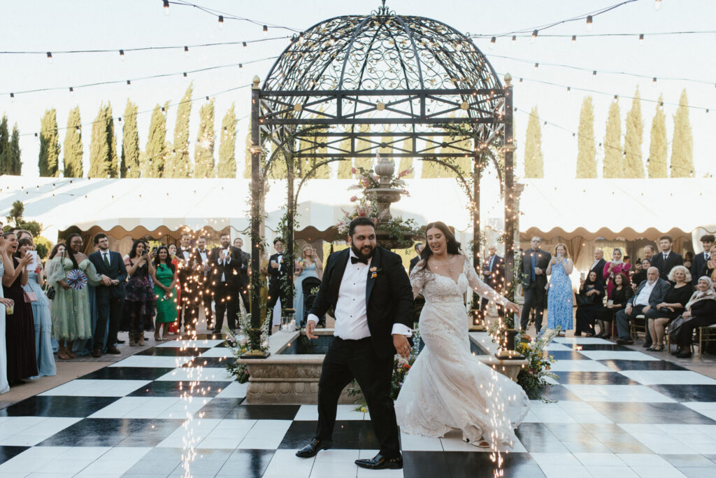 A bride and groom dance on a checkered floor under a decorative dome, surrounded by guests. Sparks rise from the floor.