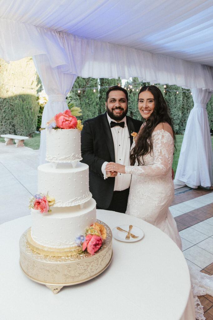 A bride and groom stand together in front of a three-tiered wedding cake with floral decorations, inside a white tent with greenery in the background.