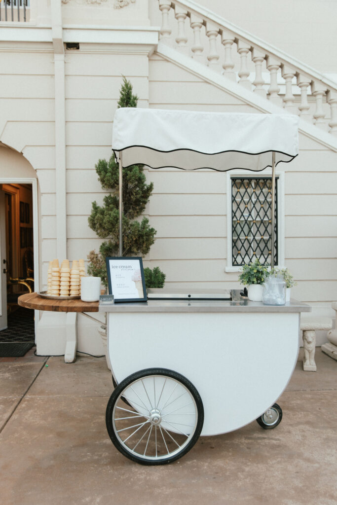 A white ice cream cart with a striped canopy is set up outdoors. Cone stacks and plant decorations are on display. A small sign is visible on the side.