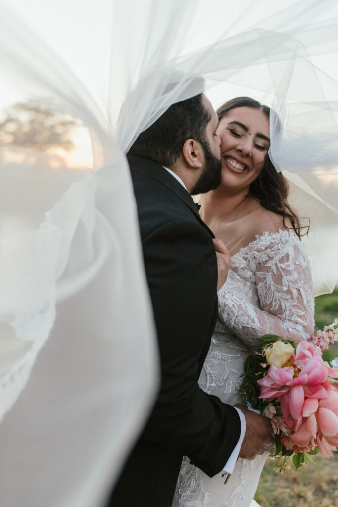 A bride and groom share a joyful moment under a flowing veil, with the bride holding pink flowers.