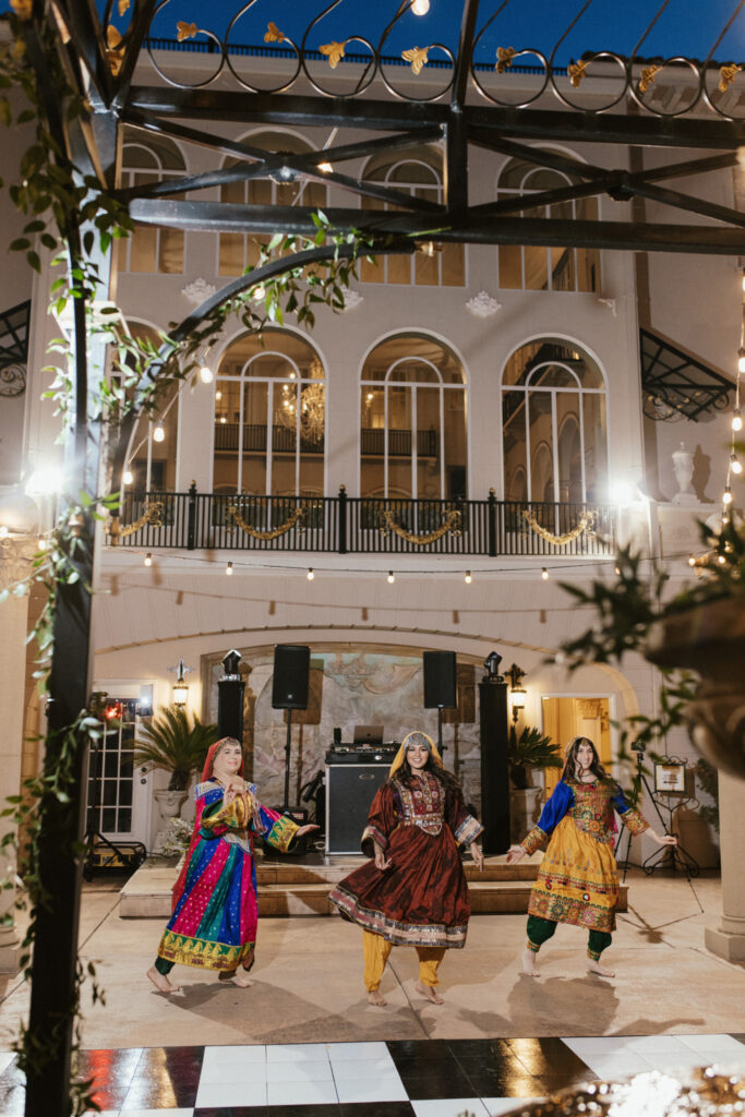 Three dancers in colorful traditional outfits perform on a checkered floor outdoors, with a grand building and decorative lights in the background.