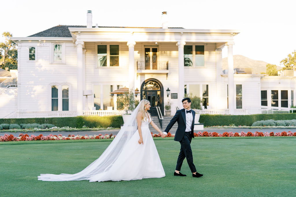 Bride in a white gown and groom in a tuxedo walk hand-in-hand on a lawn in front of a large white mansion.