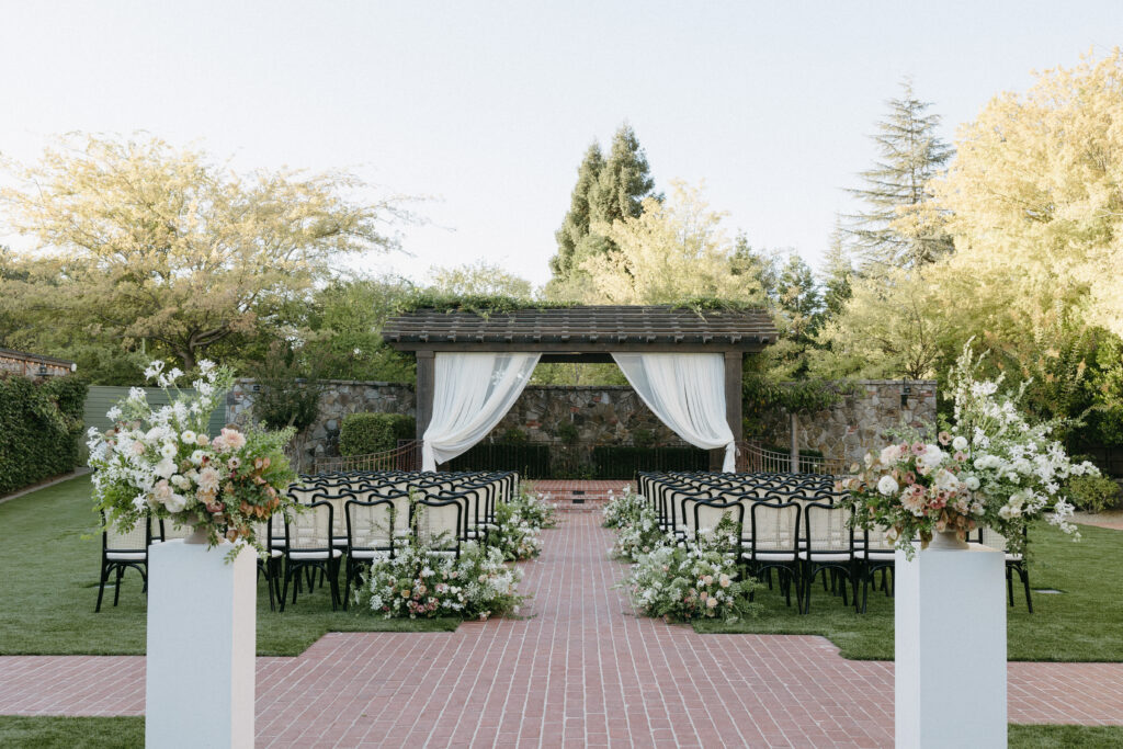 Outdoor wedding ceremony setup with chairs aligned along a brick path leading to a wood pergola with draped white fabric. Surrounded by greenery and floral arrangements.