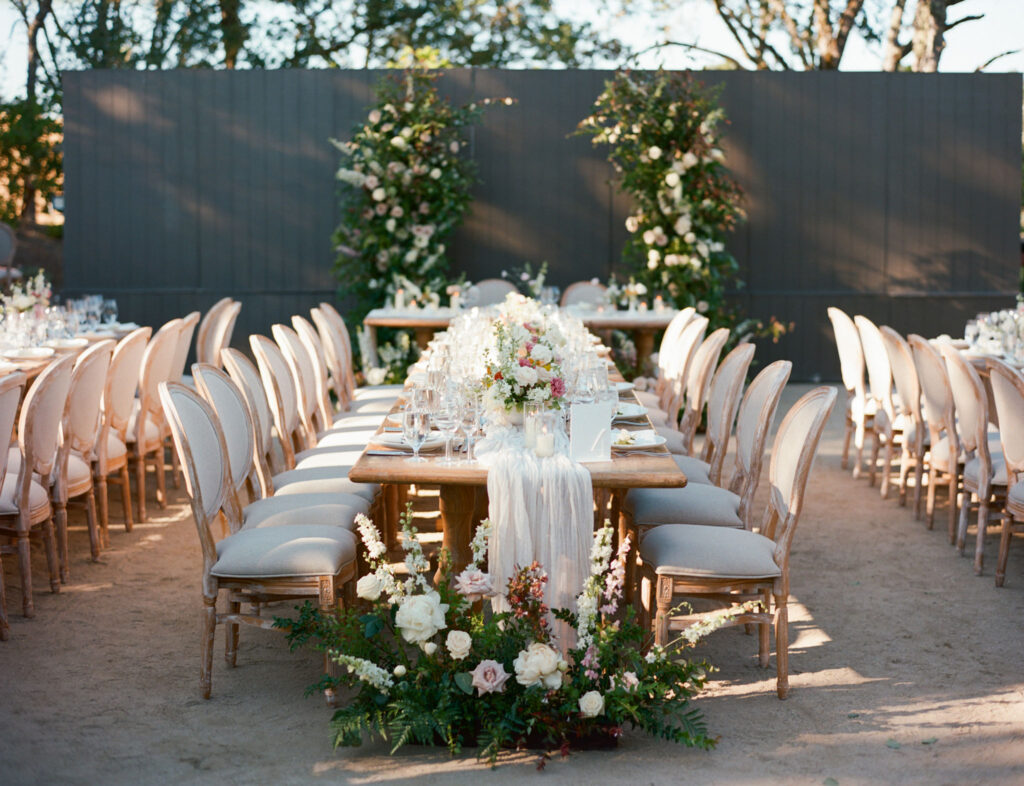 Outdoor wedding setup with two long wooden tables adorned with floral centerpieces, surrounded by white cushioned chairs. Decorative greenery and flowers in the background.