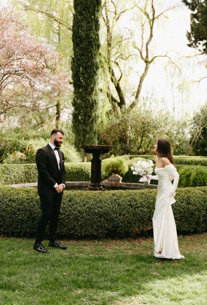 A bride and groom stand facing each other outdoors, surrounded by greenery and a hedge, with a fountain in the background. The bride holds a bouquet.