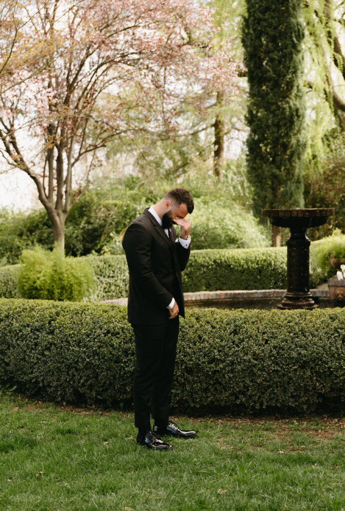 A person in a suit stands in a garden, wiping their eye. There is a fountain and trimmed hedges in the background.