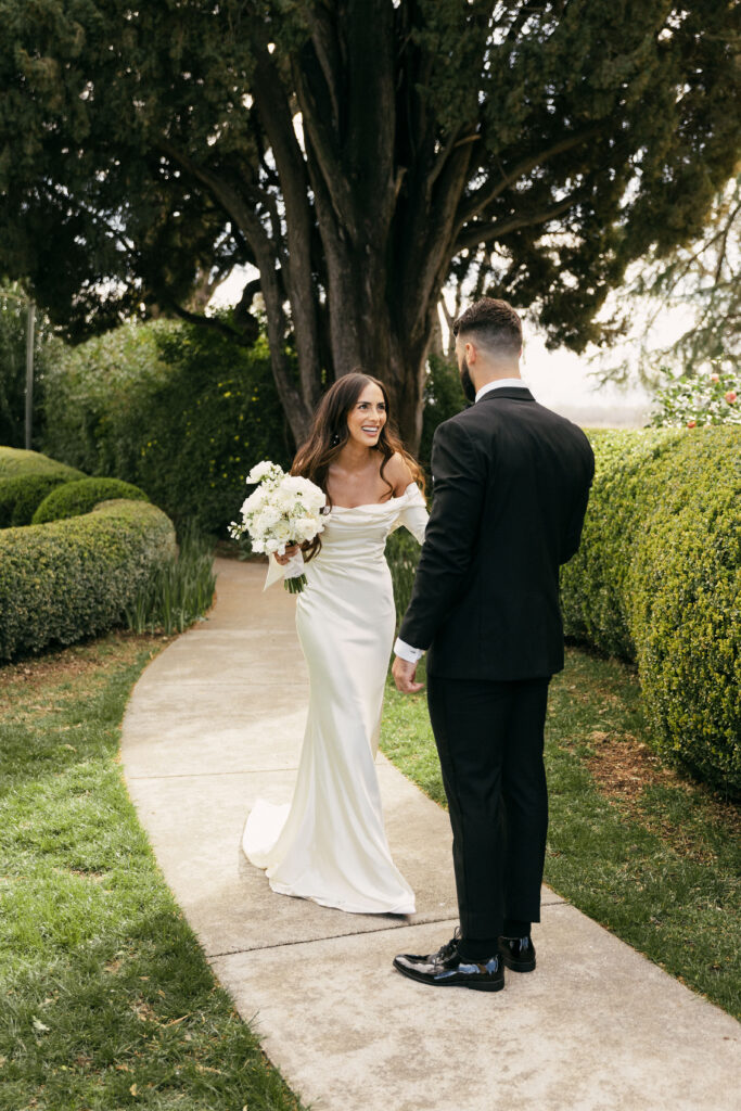 A bride in a white dress holds a bouquet, smiling at a groom in a black suit on a garden pathway. Trees and manicured bushes surround them.