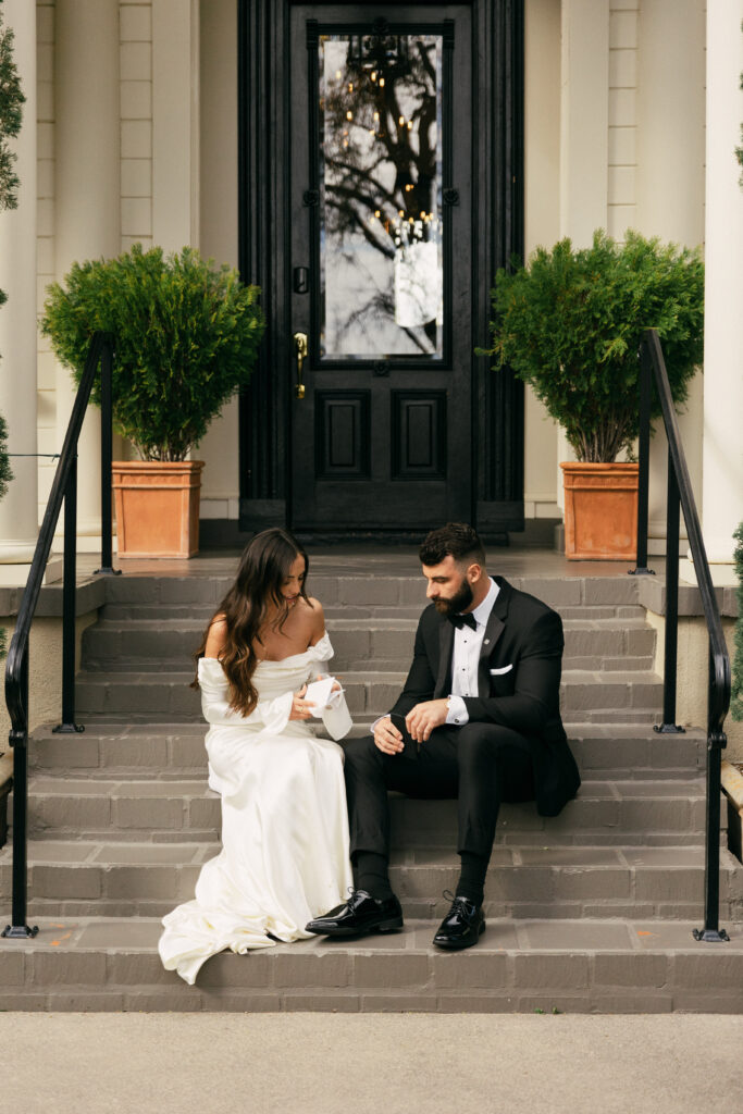 A bride and groom sit on outdoor steps in front of a black door. She wears a white gown, and he wears a black suit. Green potted plants are beside them.