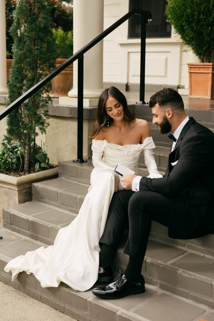 A bride in a white dress and a groom in a black tuxedo sit on steps reading from a booklet together, with plants and a building in the background.