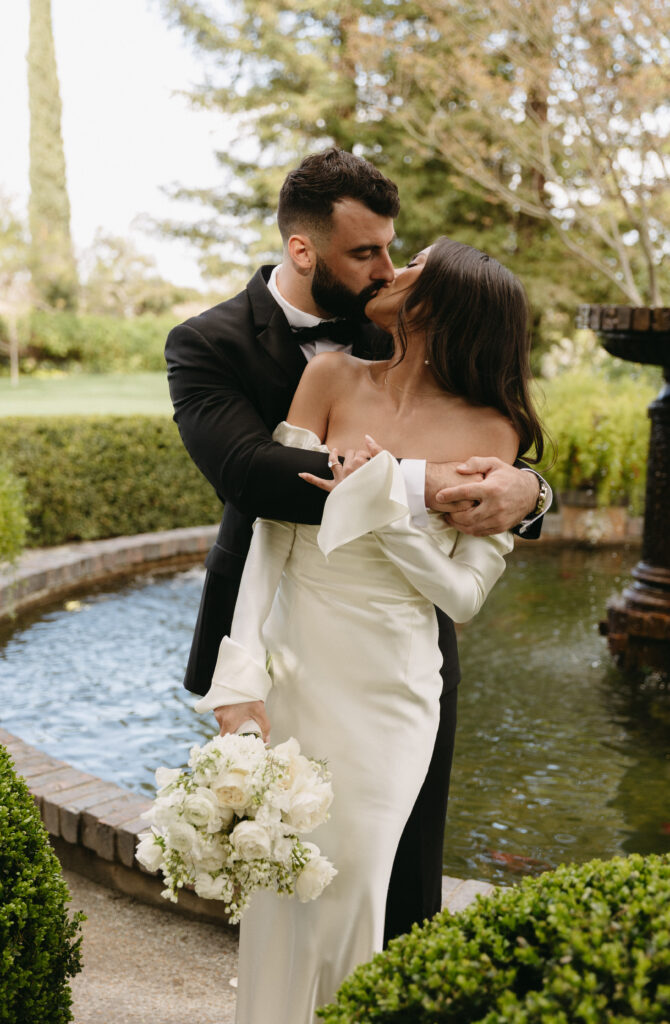 A couple in formal attire shares a kiss by a fountain in a garden. The woman holds a bouquet of white flowers.
