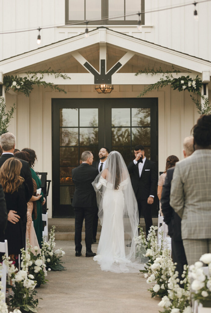 Bride and groom stand at an outdoor wedding ceremony with guests seated on either side. The bride wears a long veil. The officiant and best man are near the entrance of a building.