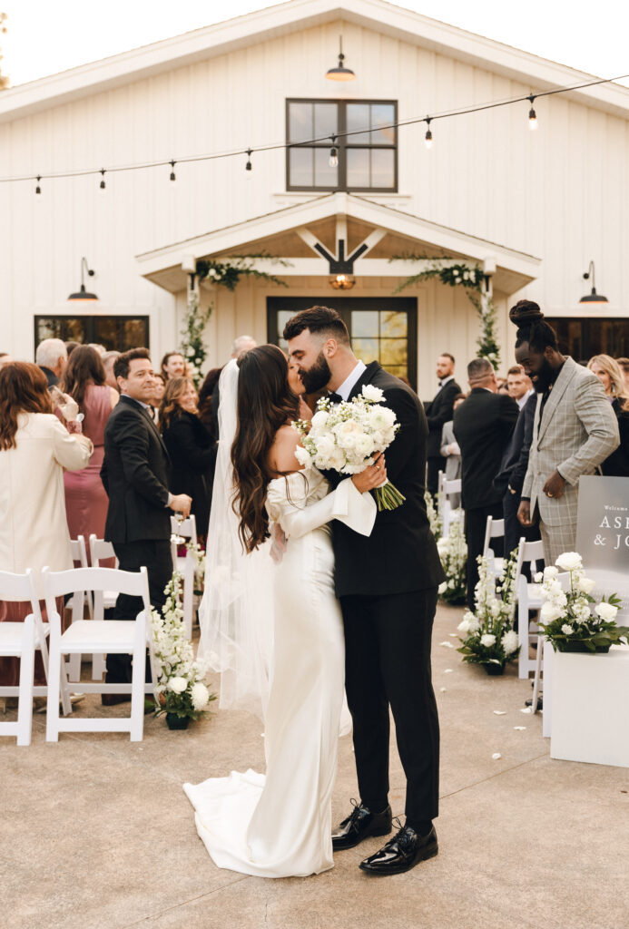 A bride and groom share a kiss in front of a white building, surrounded by guests seated in white chairs. The groom holds a bouquet, and the area is decorated with flowers and string lights.
