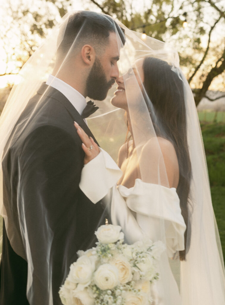 Bride and groom under a veil, smiling at each other. Bride holds a bouquet of white roses.
