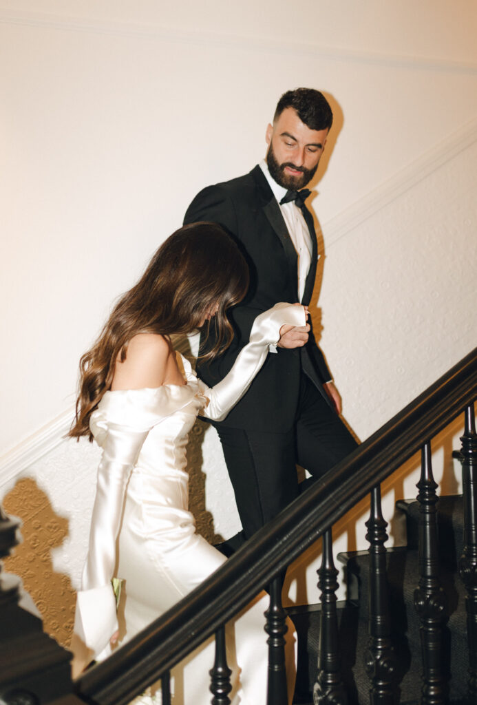 A couple in formal attire descends a staircase; the woman in a white gown and the man in a black tuxedo.