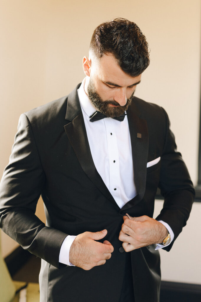 A man in a black tuxedo with a bow tie adjusts his jacket in a well-lit room.